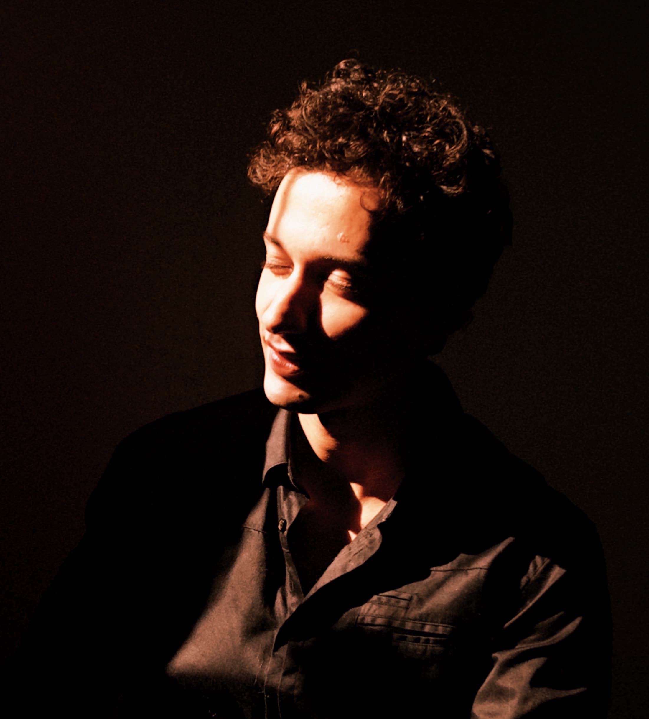 Portrait of Abdellah Ibach with curly hair in warm, low light, looking down and to the side